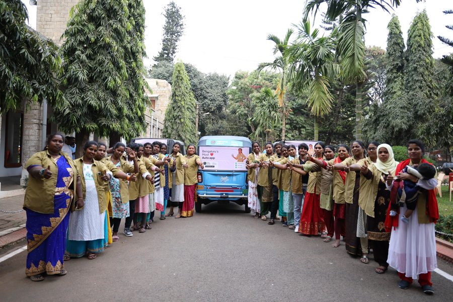 A large group of Mahila Shakti drivers giving a thumbs up around a Namma Yatri auto
